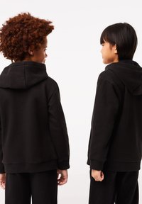 Felpe con cappuccio nere dal tessuto morbido, con polsini a coste e una vestibilità rilassata, indossate da due bambini che si fronteggiano da dietro.