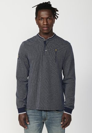 Navy long-sleeve henley shirt with textured fabric, prominent collar, a button placket, and logo accent on the chest. Paired with blue jeans.