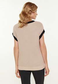 Könnyű bézs színű, fekete részletekkel ellátott, rövid ujjú kötött pulóver, amely texturált szövésű és bordázott aljjal rendelkezik. A hátsó része nyitott kialakítású.