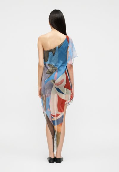 Γυναίκα γυρισμένη πλάτη που φοράει ασύμμετρο διάφανο φόρεμα με ένα ώμο, με αφηρημένα πολύχρωμα σχέδια και μαύρα επίπεδα παπούτσια.