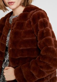 Veste en fausse fourrure marron avec un design matelassé et bouffant. Elle est dotée de boutons ronds et d'une texture douce. Le col est arrondi et les manches sont longues.