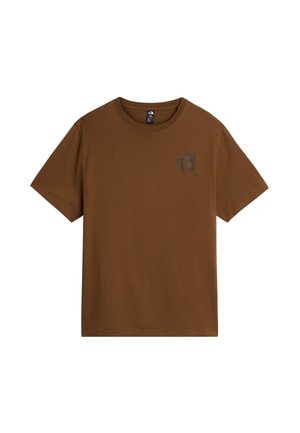 Ung mand med sort hår iført olivengrøn t-shirt og bukser, solbriller på hovedet, står med hænderne i lommerne mod en hvid baggrund.