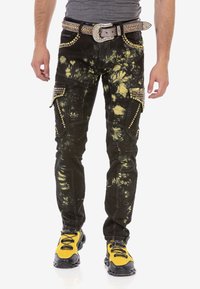 Mężczyzna w czarnych i żółtych spodni cargo w tie-dye z złotymi ćwiekami, dużą klamrą z rhinestone oraz jaskrawych żółto-czarnych sneakersach.