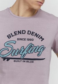 Lys lilla bomulls T-skjorte med en svart og lyseblå grafisk tekst som leser "BLEND DENIM Surfing Built in Blue" i en uformell font.