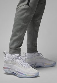 Stopy w białych sportowych trampkach z iryzującymi detalami i jasnoszarych dresach na jednolitym szarym tle.