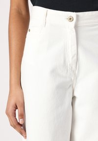 Pantalones de mezclilla blancos de talle alto con cierre de botón frontal, ligera textura y un bolsillo lateral, mostrados de la cintura hacia abajo.