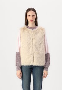 Gilet en fausse fourrure beige avec encolure ronde, texture douce et fermeture à pression sur le devant, superposé à un pull en tricot multicolore.