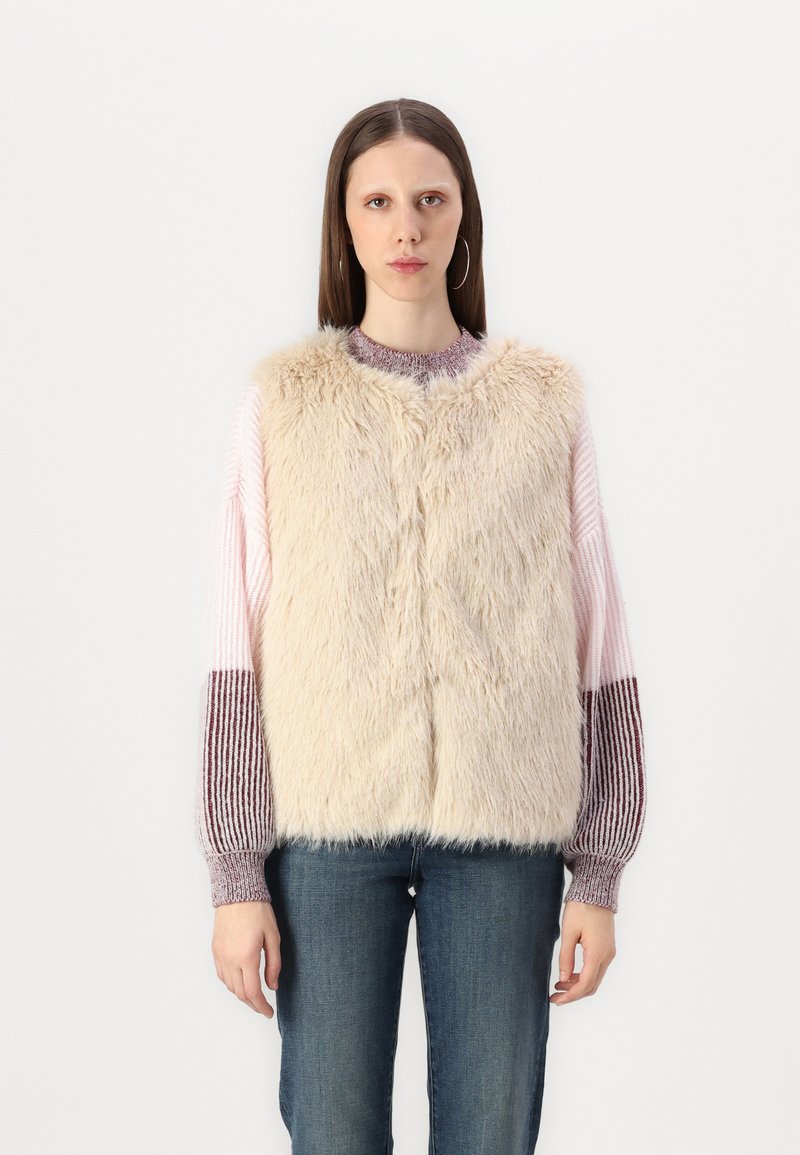 Gilet en fausse fourrure beige avec encolure ronde, texture douce et fermeture à pression sur le devant, superposé à un pull en tricot multicolore.