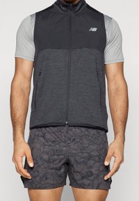 Man wearing black zip-up sleeveless vest with New Balance logo, light gray short-sleeve shirt, and black patterned athletic shorts.