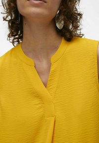 Top giallo senza maniche con scollo a V, realizzato in tessuto testurizzato, caratterizzato da motivi ondulati sottili e un design pulito e minimalista.