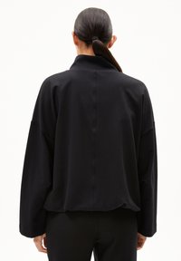 Frau mit einem dunklen Pferdeschwanz, die ein lockeres schwarzes Langarmoberteil und schwarze Hosen trägt, mit dem Rücken zu einem schlichten weißen Hintergrund.