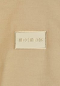Beiger Stoff mit einer glatten Textur, der ein hervorgehobenes rechteckiges cremefarbenes Etikett mit der Prägung "HECHTER" in Großbuchstaben aufweist.