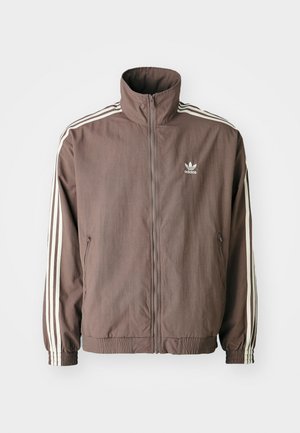 Bruine zip-up jas met een hoge kraag, voorzien van drie witte strepen op elke mouw en een geborduurd logo op de borst.