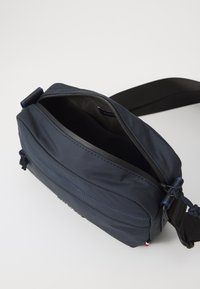 Námorná látková crossbody taška so zipsovým uzáverom, nastaviteľným popruhom a výrazným prešitím. Má hladký exteriér a vnútorné podšívanie.