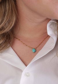 Gouden ketting met een turkoois pendant. De pendant is hartvormig met een gestructureerd oppervlak en ligt op een gesloten wit shirt.