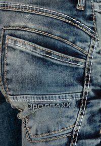 Jean en denim de couleur bleue décolorée, avec plusieurs poches, des coutures contrastées en blanc et beige, et une étiquette de marque cousue sur la poche arrière.