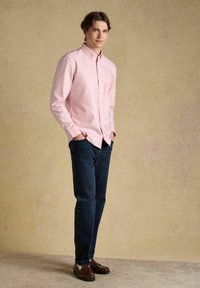 Jeune homme debout, les mains dans les poches, portant une chemise rose clair à boutons, un jean foncé et des mocassins marron, devant un fond beige.