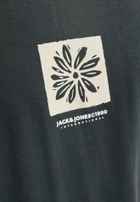 Camiseta de algodón negra con un cuadrado crema que presenta un diseño floral y el texto "JACK & JONES ©1990 INTERNACIONAL" debajo.