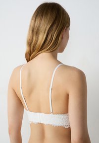 Bralette en dentelle blanche avec des bretelles réglables, un motif floral et une fermeture à crochets. Le tissu présente un design délicat et texturé.