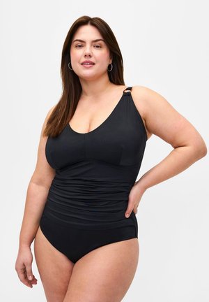 Curvy vrouw met lang bruin haar in een zwarte eenstückswimwear, staand met één hand op de heup tegen een witte achtergrond.