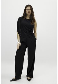 Top nero a spalla sola con tessuto drappeggiato, abbinato a pantaloni neri a vita alta. La modella indossa ballerine nere a punta e ha accessori in mano.