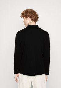 Black long-sleeve polo shirt with a collar, made of smooth fabric. Features a relaxed fit and ribbed cuffs, view from the back.