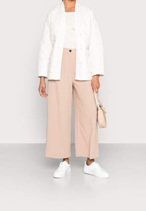 Persoon met een witte gestructureerde jas, beige brede broek, witte sneakers, en houdt een kleine beige handtas vast aan het hengsel.