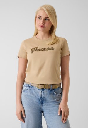 Jeune femme aux cheveux blonds porte un t-shirt beige avec le logo "Guess" et un jean bleu clair, debout devant un fond clair uni.