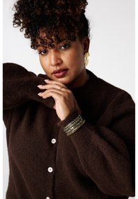 Cardigan brun et duveteux avec de gros boutons, doté d'un col arrondi et de manches bouffantes. Des bracelets métalliques dépareillés ajoutent une touche de détail.