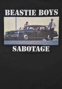Sort t-shirt med et grafisk motiv af en sort bil med to personer udenfor, teksten ovenfor lød "BEASTIE BOYS" og nedenfor "SABOTAGE" i fede hvide bogstaver.