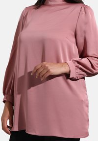 Blusa a maniche lunghe di colore rosa chiaro con colletto alto, morbido tessuto di satin e polsini arricciati. Presenta una vestibilità ampia e una caduta delicata.