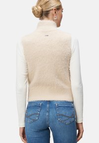 Gilet en tricot beige avec un col haut, texture douce et coupe légèrement ample. Porté par-dessus une chemise blanche à manches longues et associé à un jean bleu.
