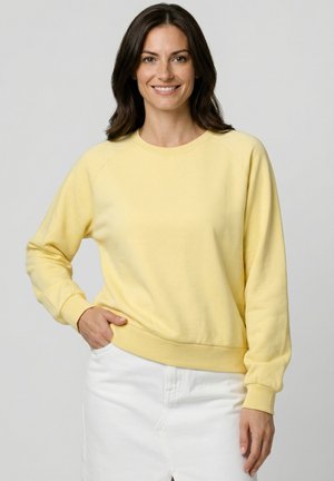 Frau mit dunklen Haaren, die lächelt, trägt ein hellgelbes Sweatshirt mit langen Ärmeln und einen weißen Rock, steht vor einem schlichten hellgrauen Hintergrund.