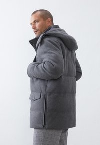 Chaqueta acolchada gris con capucha, que cuenta con un cierre frontal, bolsillos laterales y una tela texturizada. El diseño incluye una silueta estilizada.