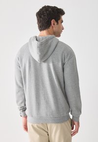 Hombre con cabello rizado oscuro, vistiendo una sudadera con capucha gris y pantalones beige, de espaldas contra un fondo claro y liso.
