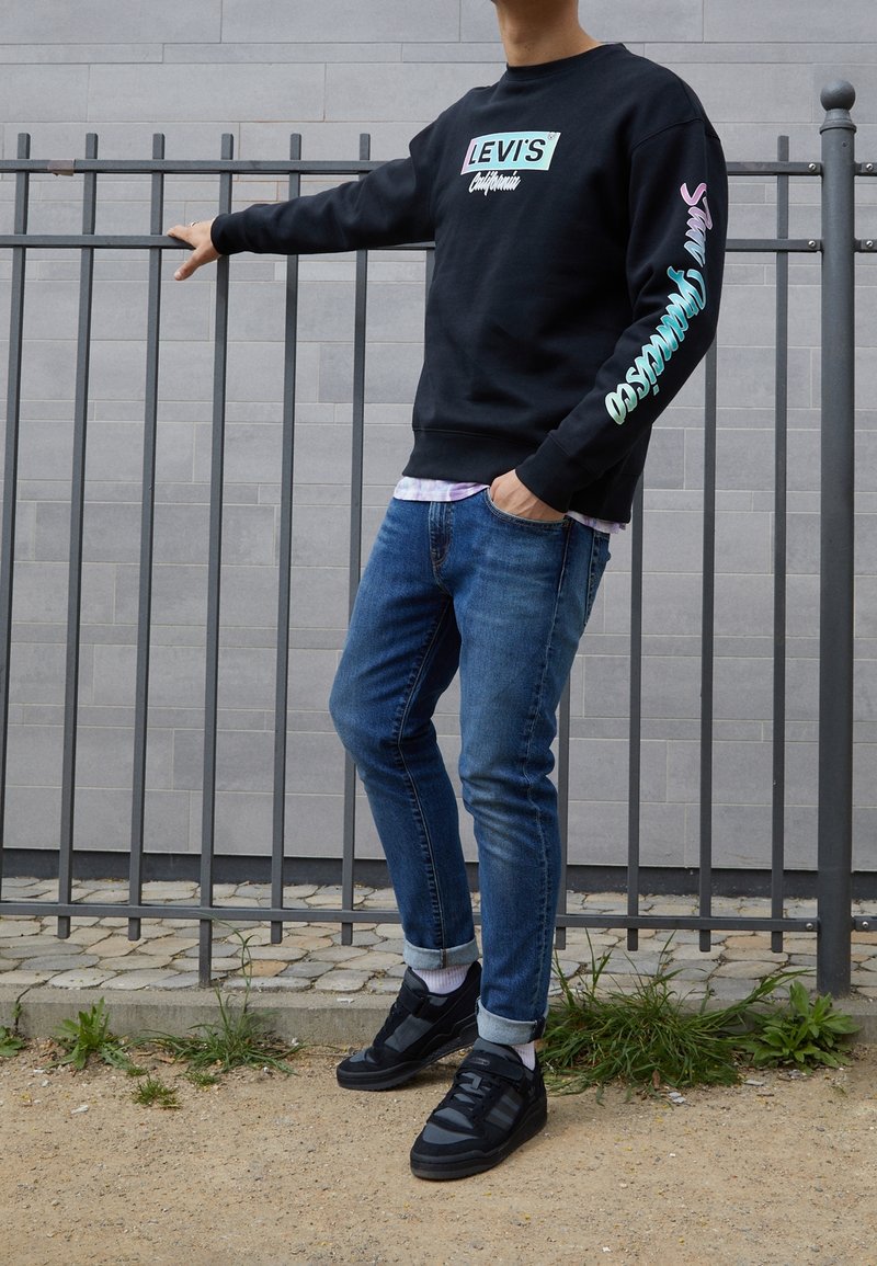 Personne portant un sweat-shirt noir Levi's, jean bleu retroussé, baskets noires, debout à l'extérieur avec une main posée sur une clôture en métal.
