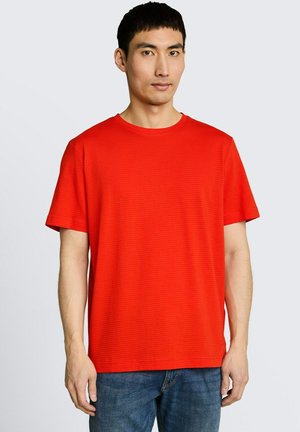 Mężczyzna stojący przodem, ubrany w zwykły, jasnoczerwony T-shirt z krótkim rękawem i niebieskie jeansy na tle jasnoszarym.