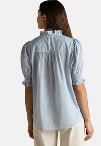 Frau, die eine hellblaue, kurzärmlige Bluse mit Rüschensaum am Kragen und an den Ärmeln trägt, steht mit dem Rücken zu einer schlichten Hintergrund.