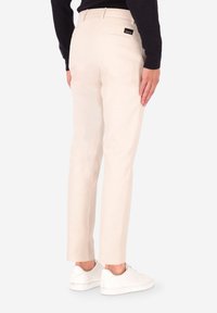 Pantaloni beige chiaro con taglio dritto, dotati di una tasca posteriore, una piccola etichetta con il marchio e indossati con sneakers bianche su uno sfondo neutro.