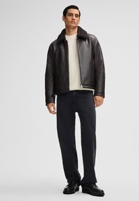 Mann steht mit einer schwarzen Lederjacke mit Lammfellkragen, einem weißen Strickpullover, schwarzen Jeans und schwarzen Schuhen vor einem einfarbigen Hintergrund.