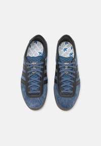 Zapatillas de ante azul con acentos de cuero negro, tres rayas negras y cordones azules. Plantilla con patrón y marca "adidas".