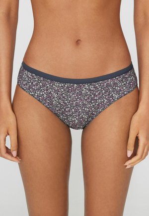 Dolní část ženského těla oblečená v tmavém květovaném spodním prádle ve stylu bikin s tmavě modrým pasem, stojící na jednobarevném pozadí.