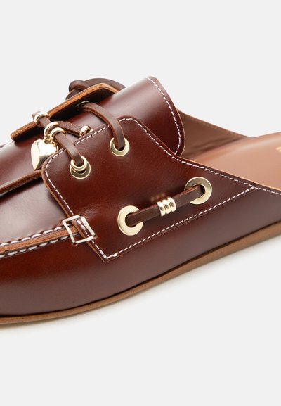 Καφέ δερμάτινο παπούτσι τύπου slip-on με χρυσά λούκια, διακοσμητική αγκράφα, λευκές ραφές και μικρό χρυσό μενταγιόν σε σχήμα καρδιάς σε δερμάτινη κορδελίτσα.