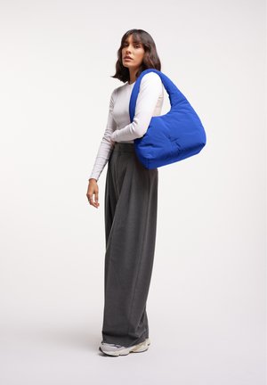 Borsa a spalla in nylon blu con texture morbida, forma oversize e manico curvo; indossata con pantaloni larghi grigi e sneakers.