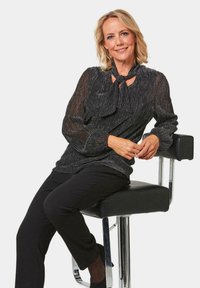 Blouse en métal noir avec un col noué, manches longues et texture transparente, associée à un pantalon noir ajusté et des talons, assise sur un tabouret de bar.