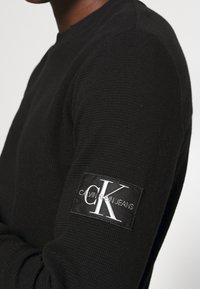 Svart långärmad sweatshirt i en texturerad tyg. Har en vävd logotyp-patch på vänster ärm med "CALVIN KLEIN JEANS" i vitt.