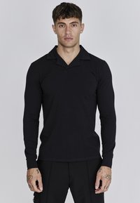 Camisa polo negra de manga larga con cuello en forma de V, textura suave y discreto logo. El corte es contemporáneo y ajustado.