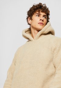Beige fleece kapucnis pulóver plüss textúrával, oversized fazonnal és nagy kapucnival. Zökkenőmentes kialakítás, látható fémrészek vagy minták nélkül.