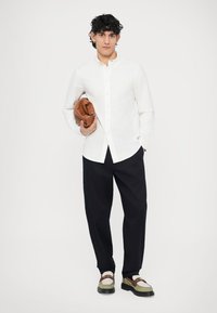 Camicia bianca con bottoni, pantaloni scuri e scarpe bicolor. Tiene in mano una borsa marrone. Sfondo chiaro, modello di fronte, capelli corti e ricci.