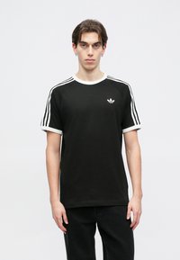 Čierne tričko s krátkym rukávom a bielym rebrovaným golierom a manžetami. Má tri biele prúžky na pleciach a malé biele logo na hrudi.